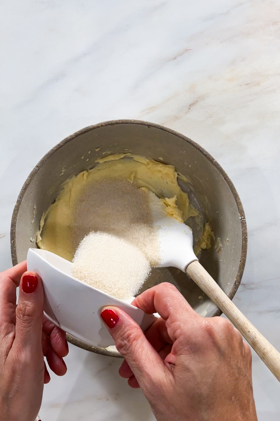 Una persona con las uñas pintadas de rojo vierte azúcar granulada de un pequeño plato blanco en un cuenco con crema de mantequilla, preparando galletas mexicanas. Una espátula blanca con mango de madera descansa sobre la encimera de mármol.