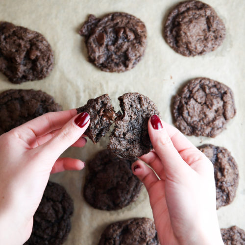 Unas manos con esmalte de uñas rojo parten una galleta de chocolate por la mitad sobre una bandeja para hornear cubierta con papel de pergamino. La bandeja contiene varias galletas de doble chocolate, dispuestas en filas. Las galletas tienen una apariencia texturizada y desmenuzable.