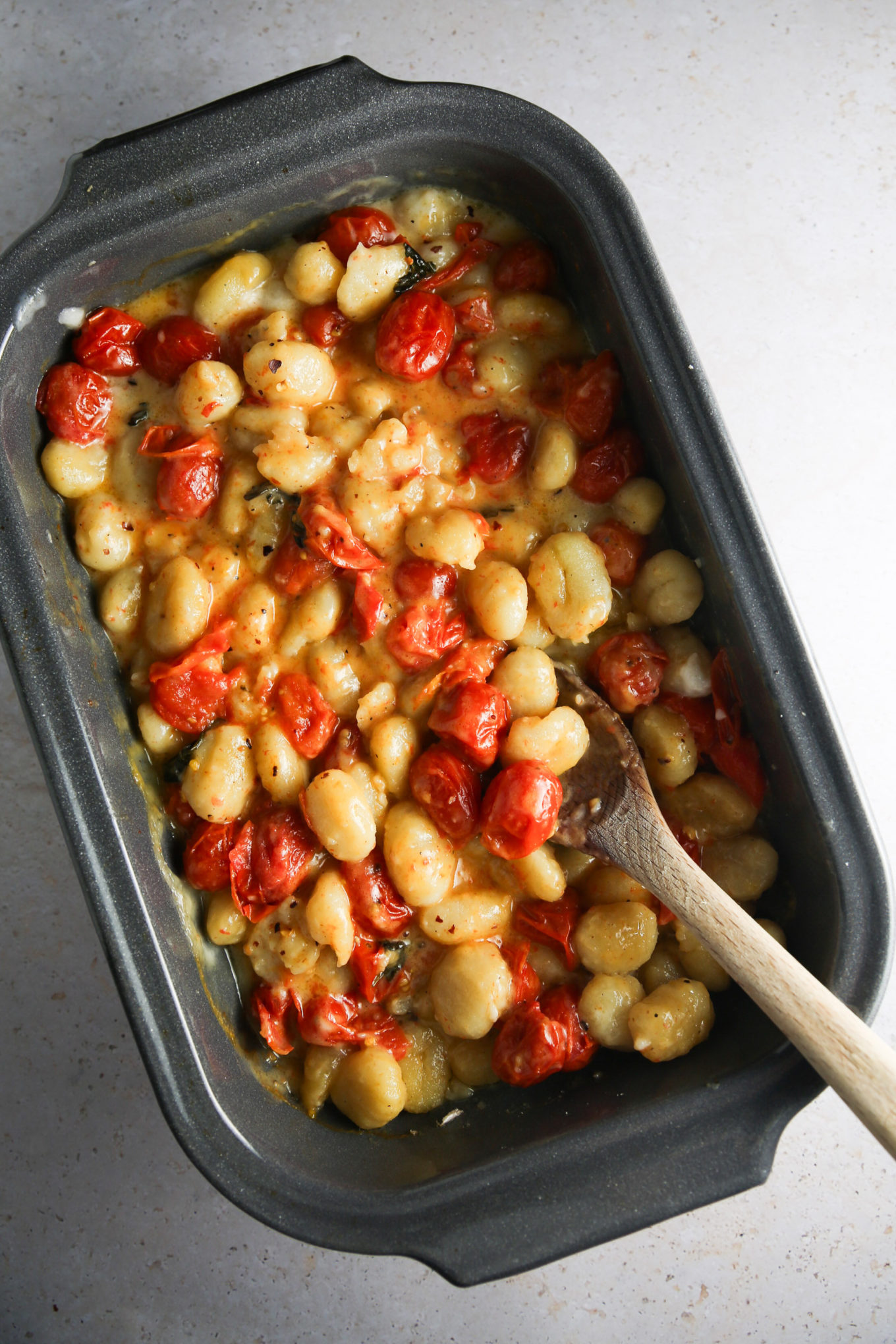 Una fuente para hornear llena de ñoquis horneados, queso fundido, queso feta y tomate cherry asado, todo mezclado con una cuchara de madera. Los ñoquis dorados y los tomates crujientes están rodeados de una salsa cremosa de queso sobre una encimera de color claro.