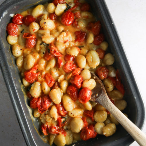 Una fuente para hornear llena de ñoquis horneados, queso fundido, queso feta y tomate cherry asado, todo mezclado con una cuchara de madera. Los ñoquis dorados y los tomates crujientes están rodeados de una salsa cremosa de queso sobre una encimera de color claro.