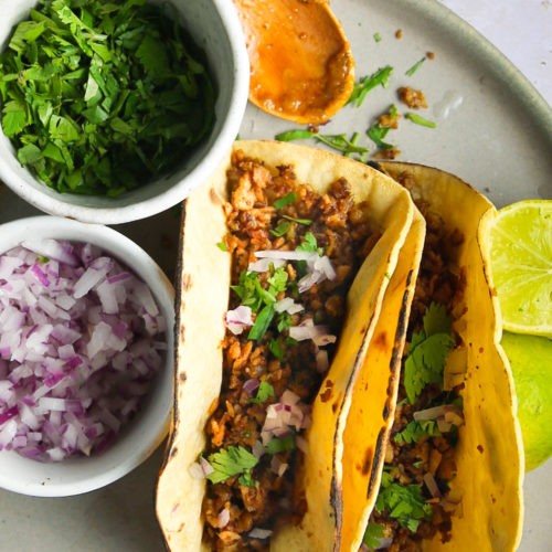 Two delicious carnitas tacos filled with seasoned meat are topped with chopped red onions and cilantro, served on a plate with lime wedges. Beside them, small bowls of fresh chopped cilantro and red onion await, with a wooden spoon resting on the edge of one bowl.