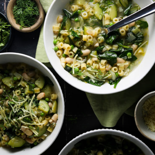 Un nutritivo plato de sopa de primavera con pasta ditalini, garbanzos, espinacas y hierbas finamente picadas, cubierto con queso rallado. La sopa se sirve en un bol blanco con una cuchara,