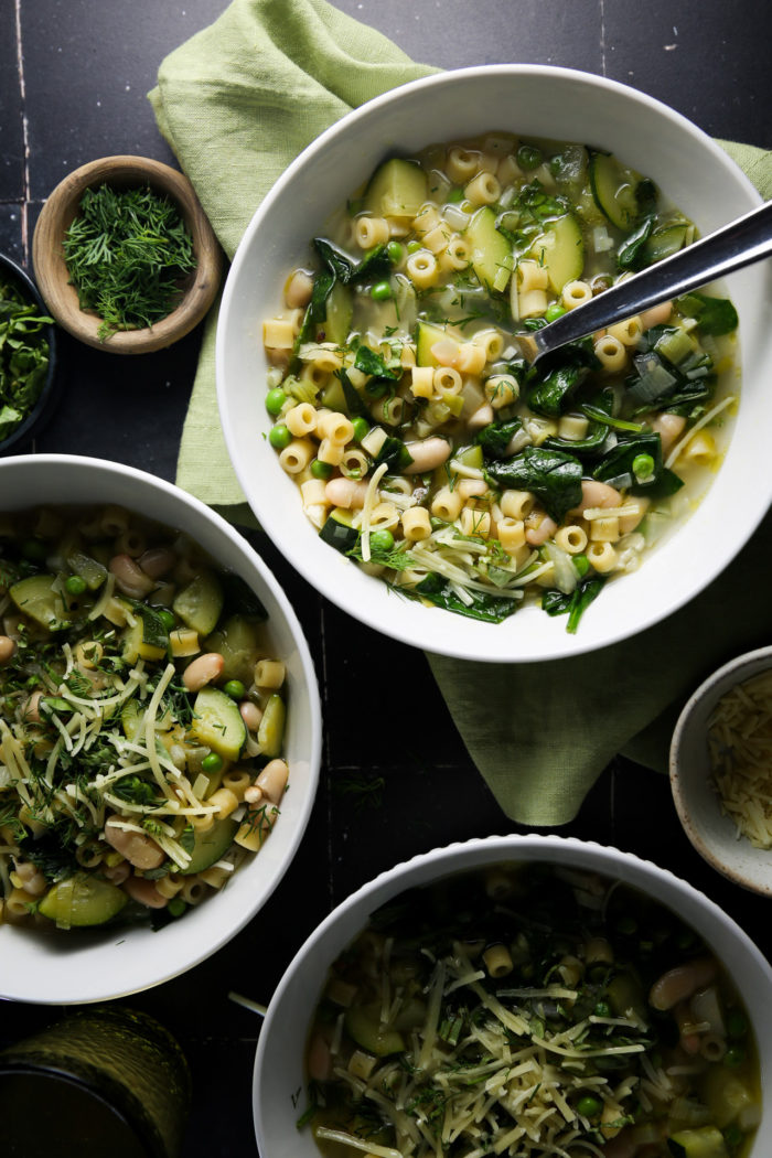 Un nutritivo plato de sopa de primavera con pasta ditalini, garbanzos, espinacas y hierbas finamente picadas, cubierto con queso rallado. La sopa se sirve en un bol blanco con una cuchara,