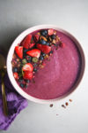 A creamy purple Smoothie Bowl is topped with fresh strawberries, blueberries, granola, and a few dried raspberries, arranged in clusters on the left side of the bowl. A gold spoon and a purple cloth napkin are placed to the left of the bowl on a light gray surface, making for an easy 5 Minute Recipe.