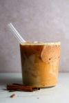 Un vaso de café helado con espuma está colocado sobre una superficie clara. En la bebida hay una pajita de cristal, y junto al vaso reposan palitos de canela. El borde está recubierto de azúcar con canela y cerca hay totopos crujientes.