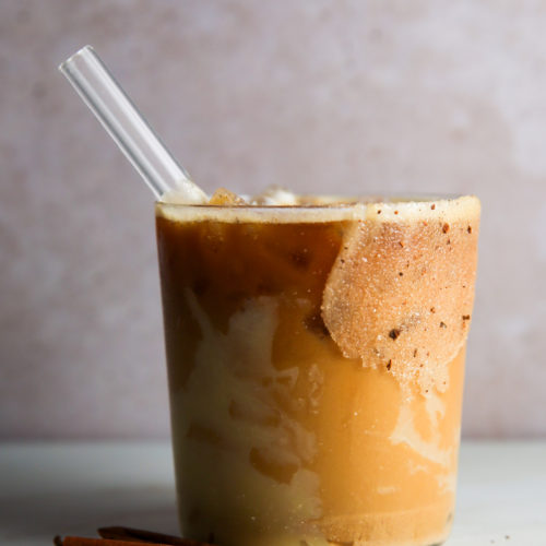 Un vaso de café helado con espuma está colocado sobre una superficie clara. En la bebida hay una pajita de cristal, y junto al vaso reposan palitos de canela. El borde está recubierto de azúcar con canela y cerca hay totopos crujientes.