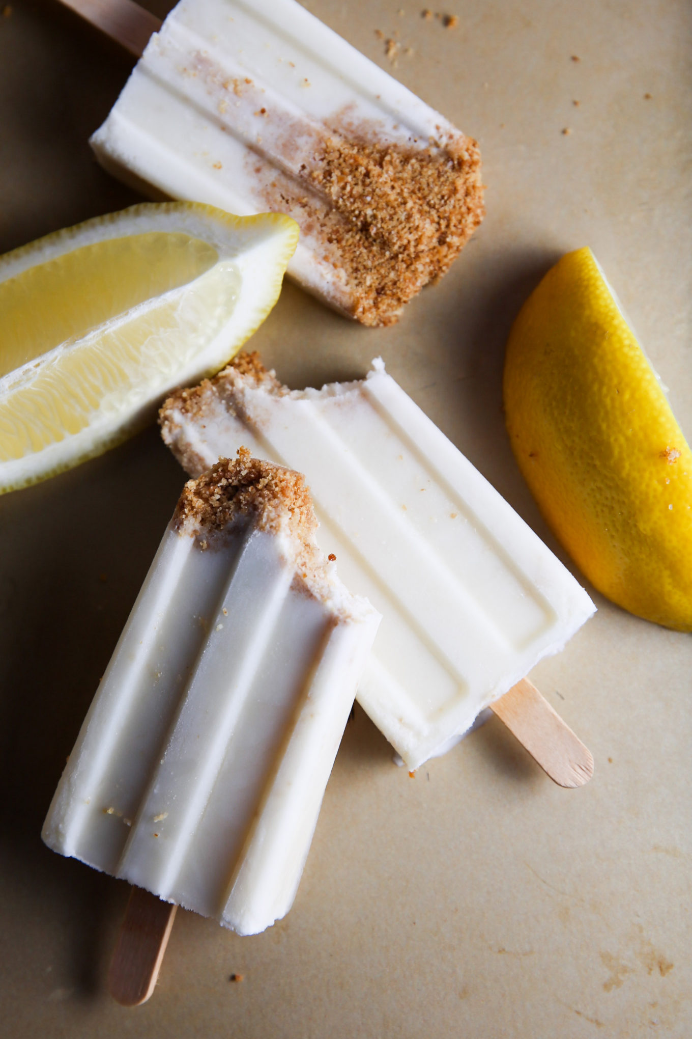 Tres paletas con sabor a tarta de limón con corteza de azúcar moreno sobre una superficie beige, acompañadas de gajos de limón fresco. Las paletas son parcialmente visibles y descansan verticalmente.