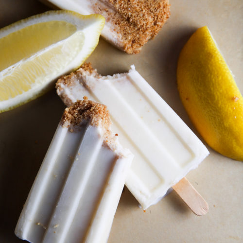 Tres paletas con sabor a tarta de limón con corteza de azúcar moreno sobre una superficie beige, acompañadas de gajos de limón fresco. Las paletas son parcialmente visibles y descansan verticalmente.