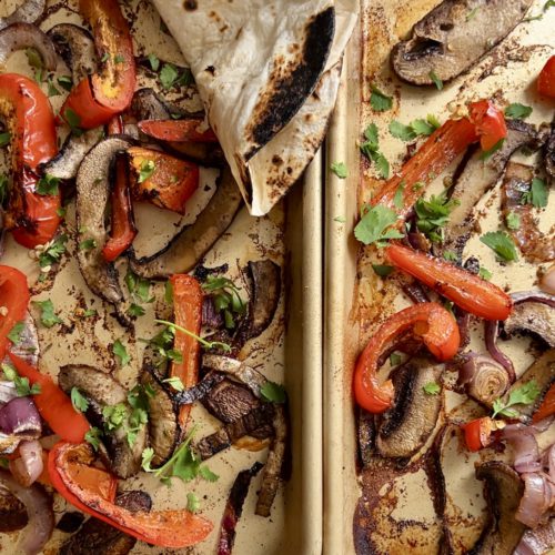 Dos sartenes llenas de verduras asadas, incluidos pimientos rojos, cebollas rojas y champiñones portobello. Las verduras sazonadas se adornan con cilantro fresco. En la esquina superior izquierda se coloca una tortilla de harina ligeramente chamuscada, cubriendo parcialmente algunas verduras: perfecto para una receta de fajitas.