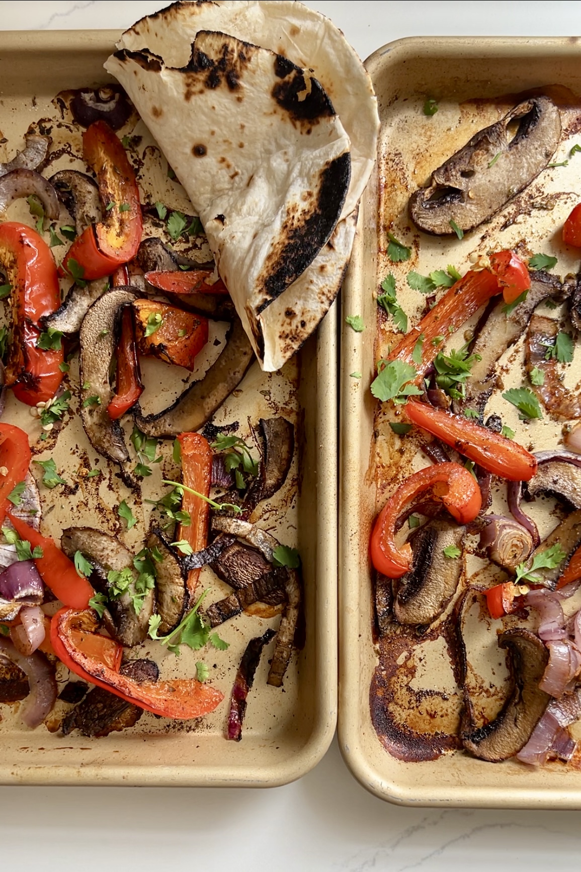 Dos sartenes llenas de verduras asadas, incluidos pimientos rojos, cebollas rojas y champiñones portobello. Las verduras sazonadas se adornan con cilantro fresco. En la esquina superior izquierda se coloca una tortilla de harina ligeramente chamuscada, cubriendo parcialmente algunas verduras: perfecto para una receta de fajitas.