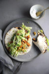 Two soft tacos filled with roasted vegetables, shredded lettuce, and crumbled cheese are placed on a gray ceramic plate. A small bowl with extra cheese and a golden spoon sits nearby. A piece of folded gray cloth is partially seen under the plate on the muted gray surface. Try this healthy recipe for sweet potato tacos!