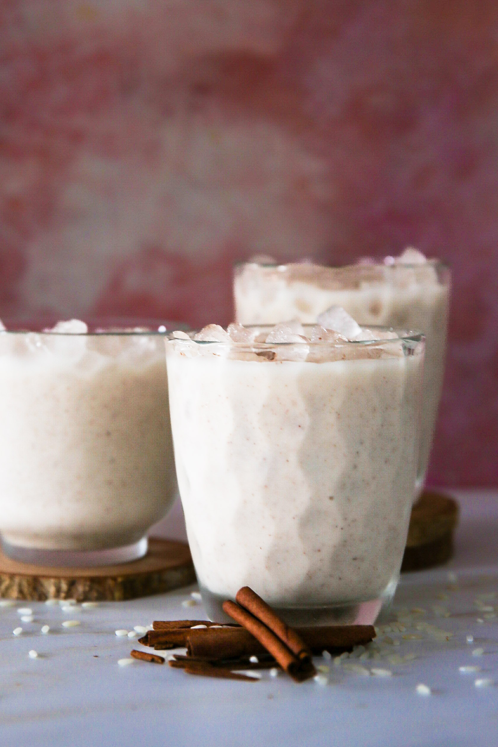 Tres vasos de cremosa horchata vegana con cubitos de hielo, colocados sobre posavasos de madera. Las bebidas se presentan sobre un fondo rosa suave, adornadas con palitos de canela y granos de arroz esparcidos en el