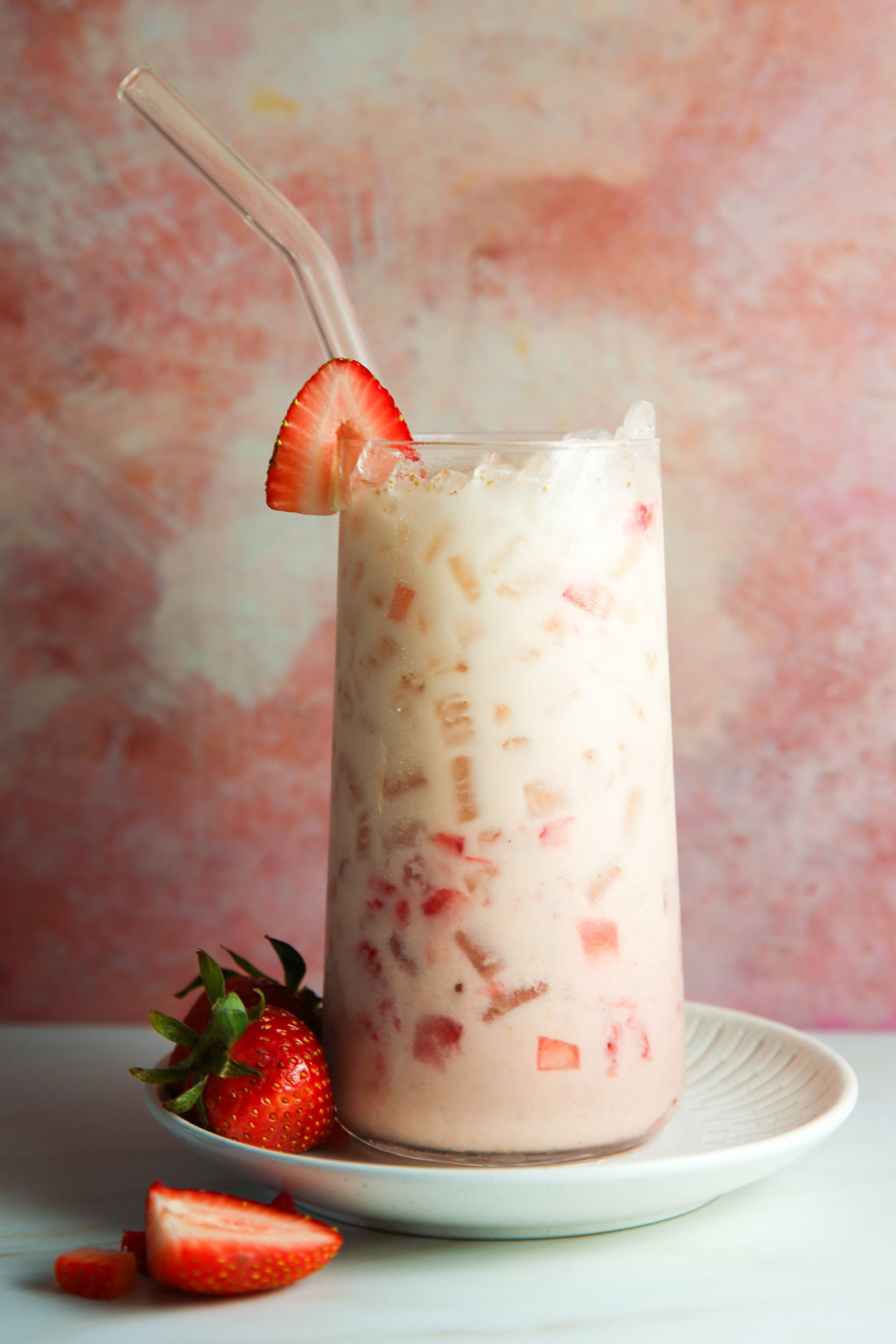 Una refrescante horchata de fresa en un vaso transparente, lleno de hielo y fresas troceadas, coronado con una fresa entera en el borde. La bebida se sirve con una pajita de cristal, en un plato ligero