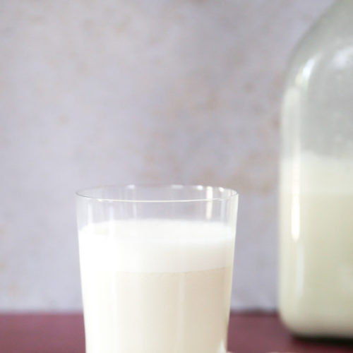 Un vaso transparente lleno de cremosa leche de anacardos descansa sobre un posavasos de madera. Se colocan tres anacardos junto al vaso. Al fondo, una gran botella de cristal que contiene más leche de anacardo está ligeramente desenfocada. Bajo el vaso hay una servilleta blanca doblada, todo sobre una superficie marrón. ¡Prepara leche de anacardos de la forma más fácil y disfruta de esta deliciosa bebida!