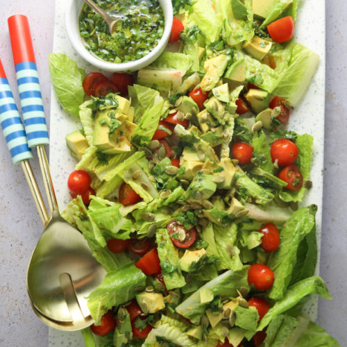 Se sirve una ensalada vibrante en un plato blanco rectangular. La ensalada lleva lechuga romana picada, tomates cherry partidos por la mitad y trozos de aguacate, adornados con semillas. En la parte superior izquierda hay un pequeño cuenco con aderezo de cilantro y, a su lado, dos cucharas con mango a rayas azules y blancas.
