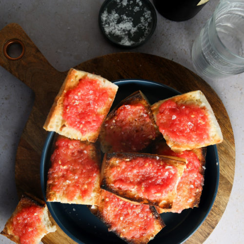 Una tabla redonda de madera para servir sostiene un plato negro cubierto con varias rebanadas de pan tostado untadas con una mezcla de tomate rojo con trozos, que recuerda al Pan Con Tomate. Al lado del plato, sobre la superficie clara de la mesa, hay un vaso de agua, una botella de vino y un cuenco pequeño de sal gruesa.