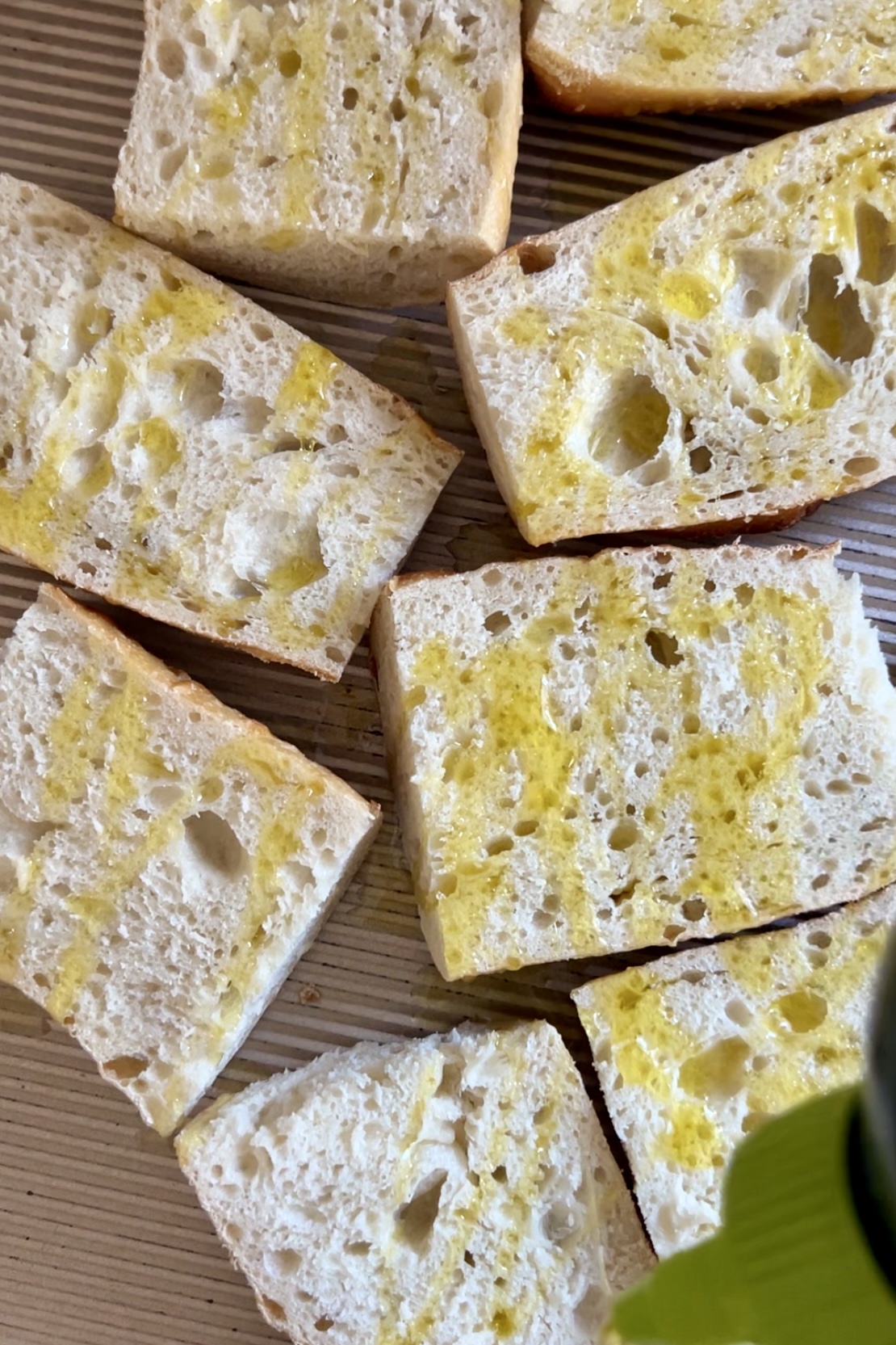 Primer plano de varias rebanadas de pan de chapata sobre una superficie de madera, perfectas para el Pan Con Tomate. El pan se rocía ligeramente con aceite de oliva, lo que le da un tinte amarillento y realza su textura porosa y aireada. Los agujeros irregulares y el aspecto suave y rústico son característicos de la chapata.