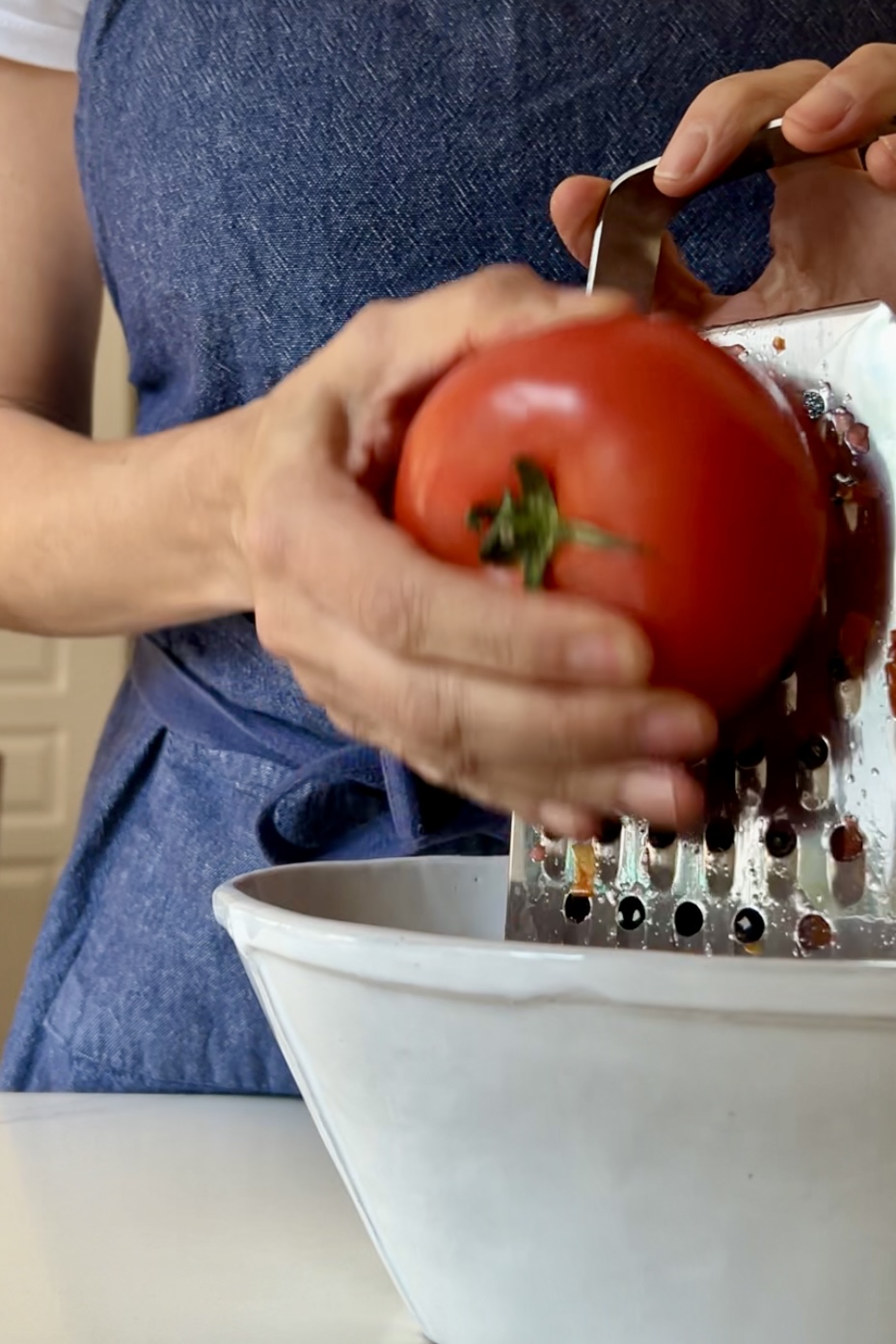 Primer plano de una persona con un delantal azul rallando un tomate rojo sobre un recipiente blanco, perfecto para preparar Pan Con Tomate. La persona sujeta el tomate con la mano izquierda mientras lo presiona contra el rallador con la derecha. La imagen se centra en las manos, el tomate y el rallador, con el fondo ligeramente difuminado.