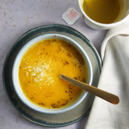 Un cuenco de sopa de pastina amarilla con una pizca de perlas de tapioca blanca, servido en un cuenco blanco sobre un plato gris verdoso con una cuchara dorada. A su lado hay una taza blanca que contiene un líquido amarillo y una etiqueta con una bolsita de té usada. A la derecha se ve parcialmente una servilleta de tela de color crema.