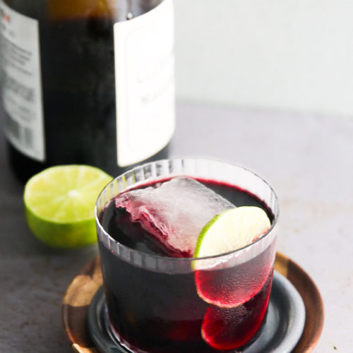 Un vaso lleno de vino tinto, adornado con una rodaja de lima y un gran cubito de hielo, se asienta sobre un posavasos de madera y cerámica. Detrás del cristal hay una botella de Tinto de Verano con una etiqueta pegada, y una lima partida por la mitad está colocada junto a la botella sobre una superficie gris claro.