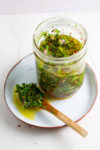 Un tarro de cristal lleno de salsa chimichurri de hierbas verdes sobre un plato blanco. Una cuchara de madera, untada en la salsa con aceite visible y hierbas frescas, descansa junto al tarro, perfecta para acompañar los totopos crujientes.
