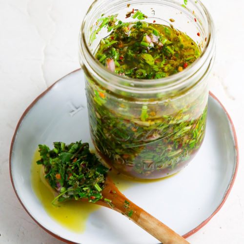 Un tarro de cristal lleno de salsa chimichurri de hierbas verdes sobre un plato blanco. Una cuchara de madera, untada en la salsa con aceite visible y hierbas frescas, descansa junto al tarro, perfecta para acompañar los totopos crujientes.