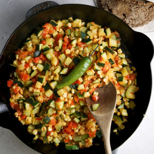 Sobre una encimera blanca hay una sartén Staub negra, llena de una colorida mezcla de verduras picadas, como calabacines, tomates y maíz.
Encima de las verduras hay un chile verde, y en la sartén hay una cuchara de madera.
Al fondo, hay un pequeño cuenco con sal y un montón de tortillas.