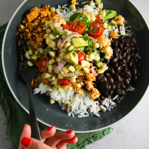 Una mano con esmalte de uñas rojo y una pulsera de oro sostiene un tenedor cerca de un cuenco de comida. El vibrante Burrito Bowl contiene arroz blanco, frijoles negros, migas de tofu sazonadas y una mezcla de ensalada fresca de tomates, aguacate, cebolla roja y maíz. El cuenco reposa sobre un paño verde contra una superficie gris claro.