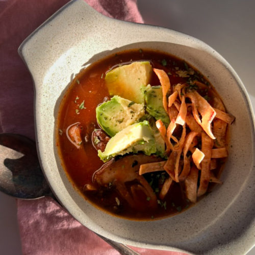 Un plato de sopa de tortilla relleno con un rico caldo rojo, cubierto con rodajas de aguacate, tiras de tortilla crujientes, pollo desmenuzado y hierbas. El cuenco reposa sobre una servilleta de tela rosa, con una cuchara metálica visible en un lateral. Esta abundante comida se parece al tradicional caldo de chipotle. El entorno está iluminado por luz natural.