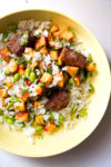 A vibrant rice bowl featuring fresh, colorful diced vegetables like edamame and bell peppers. Cubes of Mexican-spiced tofu are scattered on top, and the dish is garnished with chopped herbs. Finely mixed ingredients create an appetizing meal bursting with flavor.