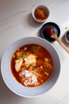 Sobre una superficie de mármol blanco hay un cuenco de sopa roja picante con tofu, arroz y cebollas verdes picadas. Cerca hay totopos crujientes, un pequeño cuenco de rábanos encurtidos y un plato de pasta de chile rojo con una cuchara de madera. La escena parece brillante y ordenada.