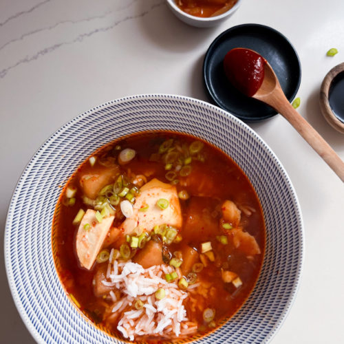 Sobre una superficie de mármol blanco hay un cuenco de sopa roja picante con tofu, arroz y cebollas verdes picadas. Cerca hay totopos crujientes, un pequeño cuenco de rábanos encurtidos y un plato de pasta de chile rojo con una cuchara de madera. La escena parece brillante y ordenada.