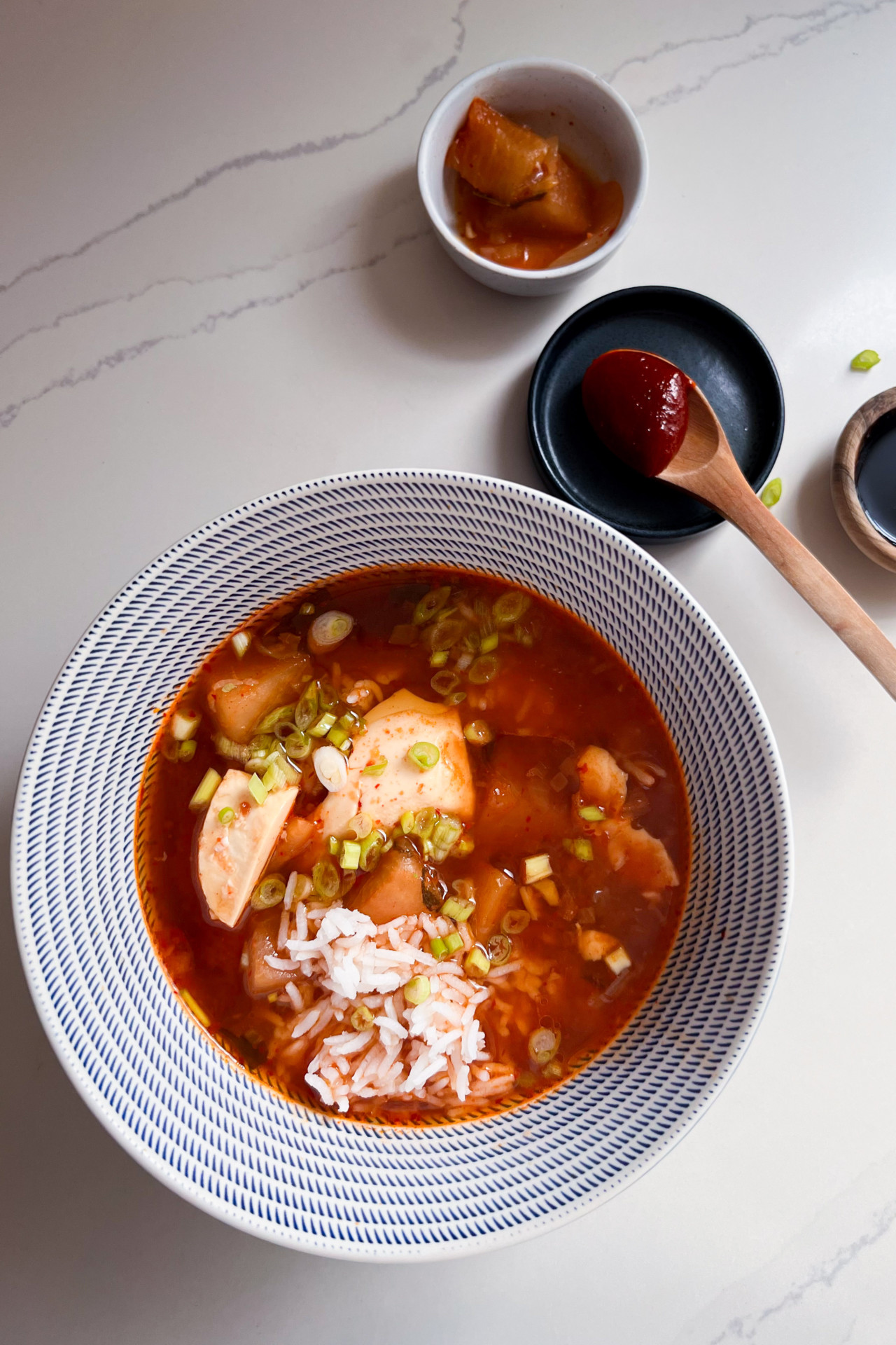 Sobre una superficie de mármol blanco hay un cuenco de sopa roja picante con tofu, arroz y cebollas verdes picadas. Cerca hay totopos crujientes, un pequeño cuenco de rábanos encurtidos y un plato de pasta de chile rojo con una cuchara de madera. La escena parece brillante y ordenada.