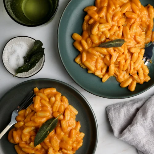 Two green plates of gnocchi in a creamy orange sauce, each garnished with a sage leaf. A green glass with a drink, a small bowl of salt, a sage leaf decoration, and a folded gray napkin are placed around them on a white marble surface.