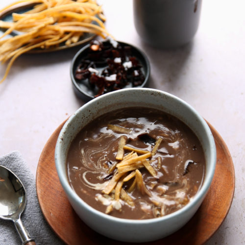 Sobre un plato de madera hay un cuenco de cremosa sopa de frijol con crujientes totopos. A su lado hay una cuchara sobre una servilleta gris, un plato pequeño de chiles rojos oscuros, tiras de tortilla extra y una jarra gris oscura, todo sobre una superficie clara.