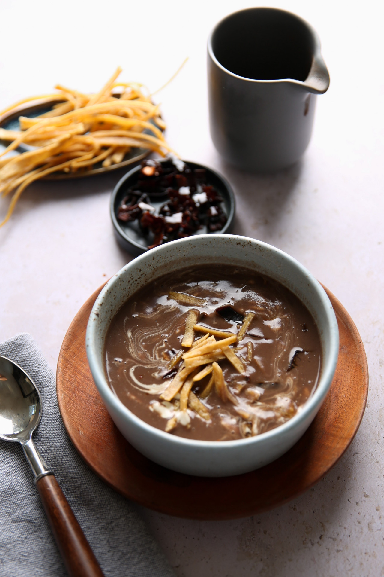 Sobre un plato de madera hay un cuenco de cremosa sopa de frijol con crujientes totopos. A su lado hay una cuchara sobre una servilleta gris, un plato pequeño de chiles rojos oscuros, tiras de tortilla extra y una jarra gris oscura, todo sobre una superficie clara.