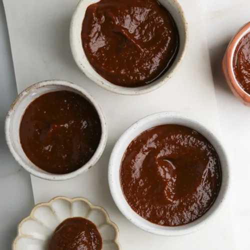 Sobre una superficie blanca se colocan cuatro cuencos pequeños llenos de una salsa espesa y marrón. Tres cuencos están hechos de cerámica, mientras que uno es un plato con forma de flor con una cuchara de madera al lado, cubierta de salsa. La textura de la salsa parece suave y espesa.