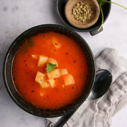 Un cuenco negro de sopa roja con dados de patata, adornado con cilantro, descansa sobre un paño gris junto a una cuchara. Encima, un pequeño plato negro contiene hierbas verdes y más cilantro, compañeros perfectos para la sopa de frijol o los totopos crujientes.