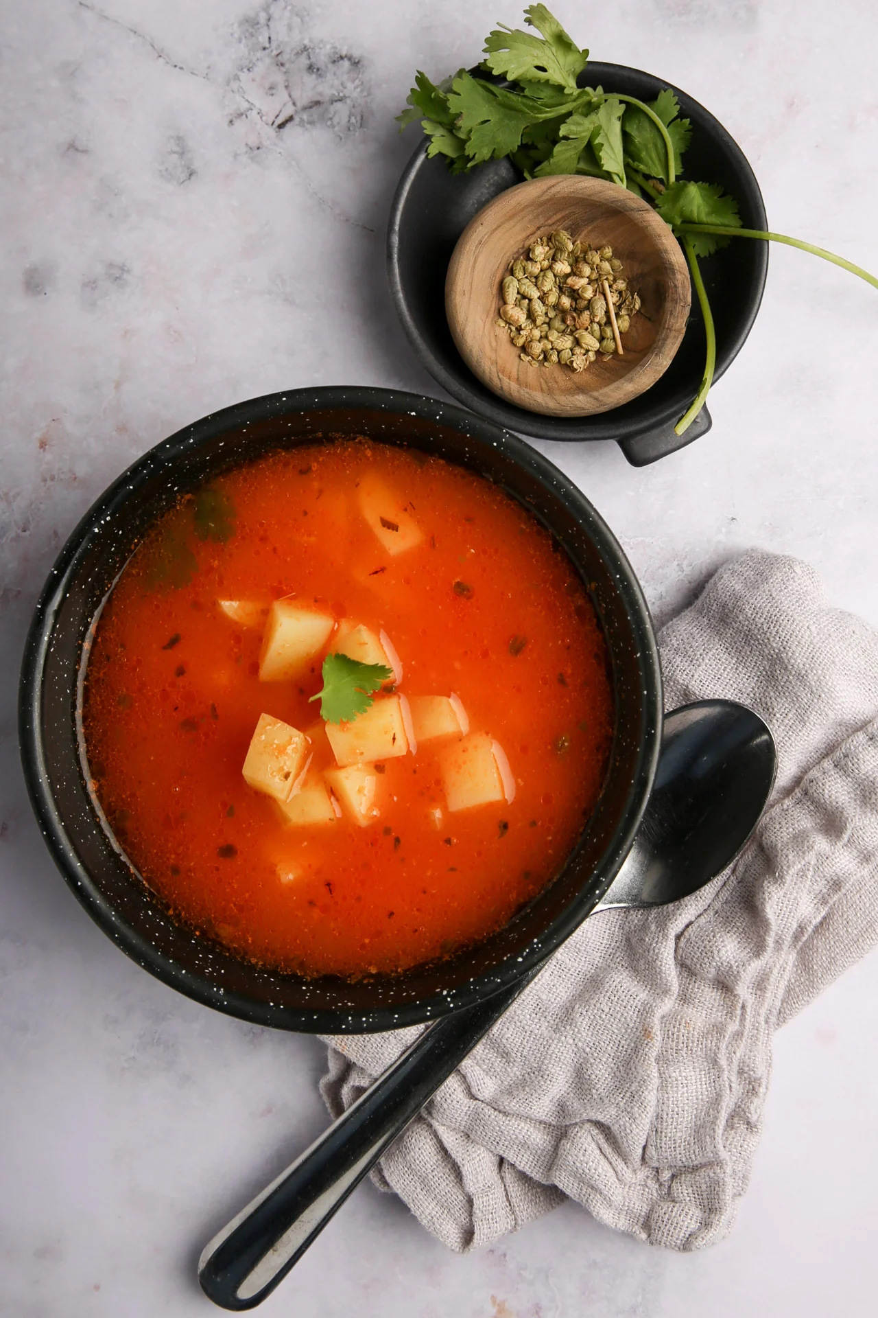 Un cuenco negro de sopa roja con dados de patata, adornado con cilantro, descansa sobre un pa&ntilde;o gris junto a una cuchara. Encima, un peque&ntilde;o plato negro contiene hierbas verdes y m&aacute;s cilantro, compa&ntilde;eros perfectos para la sopa de frijol o los totopos crujientes.