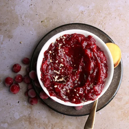 Un bowl de la salsa de arándanos más fácil de hacer, adornado con nueces picadas, descansa sobre un plato oscuro con una cuchara dorada. Los arándanos frescos esparcidos por la superficie texturizada añaden un toque rústico a esta encantadora composición.