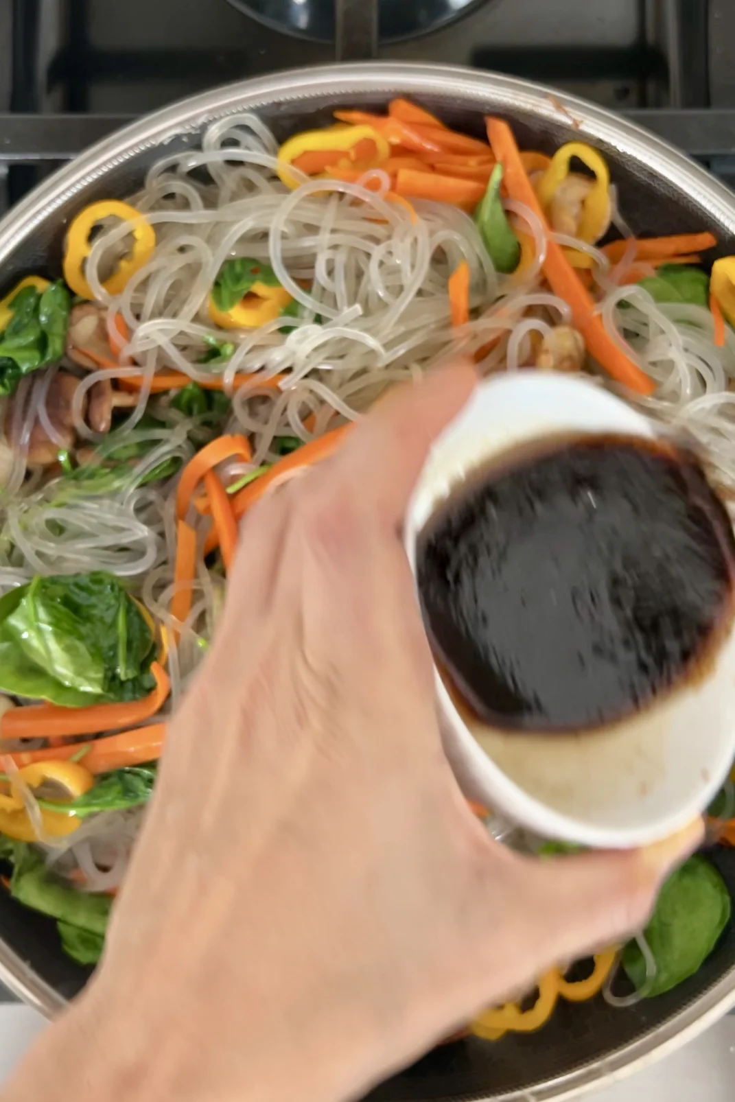 Vertiendo salsa de japchae sobre fideos de cristal y verduras en un sart&eacute;n