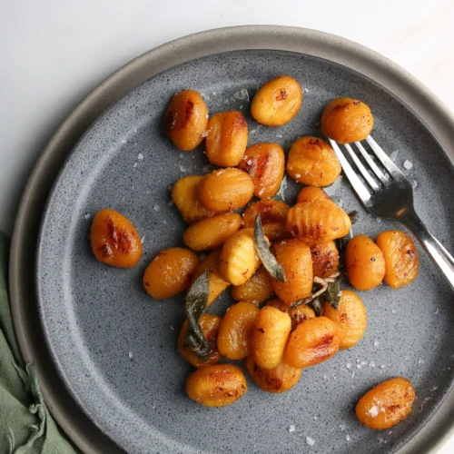 Un plato gris con ñoquis salteados, adornado con hojas de salvia crujientes y espolvoreado con escamas de sal marina. Sobre el plato hay un tenedor y, a su lado, se ve parcialmente un paño verde. Los ñoquis son de color marrón dorado y crean un contraste con el plato y el fondo.