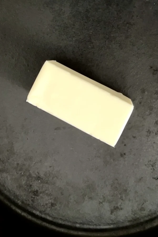 A rectangular block of butter is placed in the center of a dark, non-stick pan, ready to transform into a rich brown butter sauce. Solid and unwrapped, it rests on a clean surface, while the pan's texture peeks from the edges, anticipating the sizzle of pan-fried gnocchi.