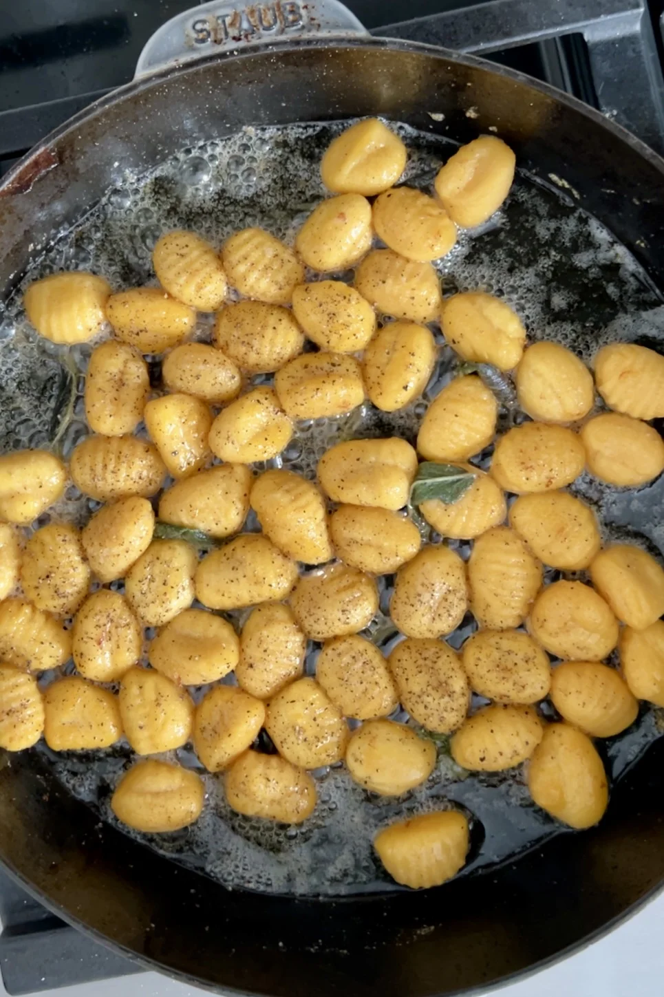 Golden pan-fried gnocchi sizzle in a black skillet with a lemon brown butter sauce and sage. The gnocchi, speckled with ground black pepper, appear perfectly seasoned. Butter bubbles around the edges, giving the dish a rich and savory look.