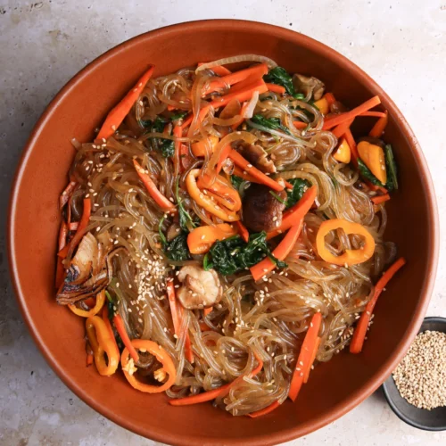Un cuenco de fideos de cristal mezclados con zanahorias cortadas en rodajas, espinacas, champiñones y pimientos morrones. El plato está adornado con semillas de sésamo y tiene un aspecto brillante y condimentado. Cerca hay un plato negro pequeño con semillas de sésamo adicionales, colocado sobre una superficie de piedra clara.