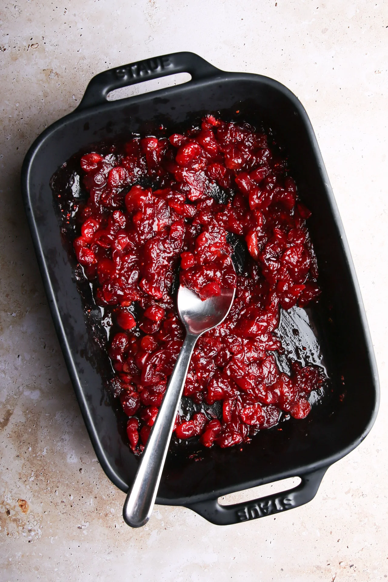 Una fuente de horno negra llena de salsa de arándanos rojos, la guarnición más fácil de preparar, brilla tentadora. En su interior reposa una cuchara de plata, con el mango elegantemente apoyado en el borde, como testimonio de haberla removido recientemente. El plato está colocado sobre una superficie clara y moteada, que contrasta con el recipiente oscuro.