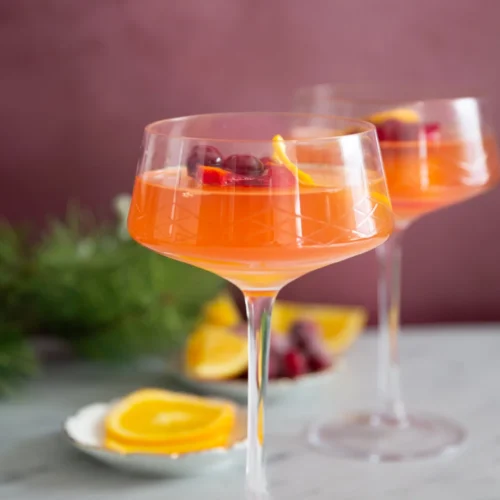 Dos copas de cóctel llenas de una bebida de color naranja intenso, decoradas con rodajas de naranja y arándanos rojos. Las copas están sobre una superficie de mármol con un fondo difuminado de naranjas en rodajas y vegetación, contra una pared de color malva suave.