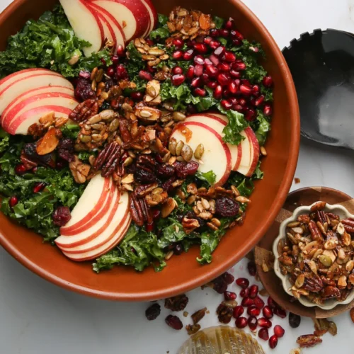 Un cuenco marrón rebosante de ensalada de col rizada está cubierto con manzanas rojas cortadas en rodajas y granos de granada, todo ello realzado con una mezcla de frutos secos. Cerca, aguardan un pequeño cuenco de frutos secos y una cuchara negra para servir. Debajo hay un vaso de vinagreta de sidra de manzana, rodeado de semillas esparcidas por la superficie blanca.