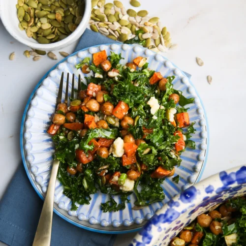 A vibrant kale salad with roasted chickpeas, crumbled cheese, and roasted sweet potatoes sits on a blue plate with a fork. Small bowls of pumpkin and sunflower seeds accompany it, all set atop a blue napkin. Try with Maple Dijon Dressing for extra flavor.