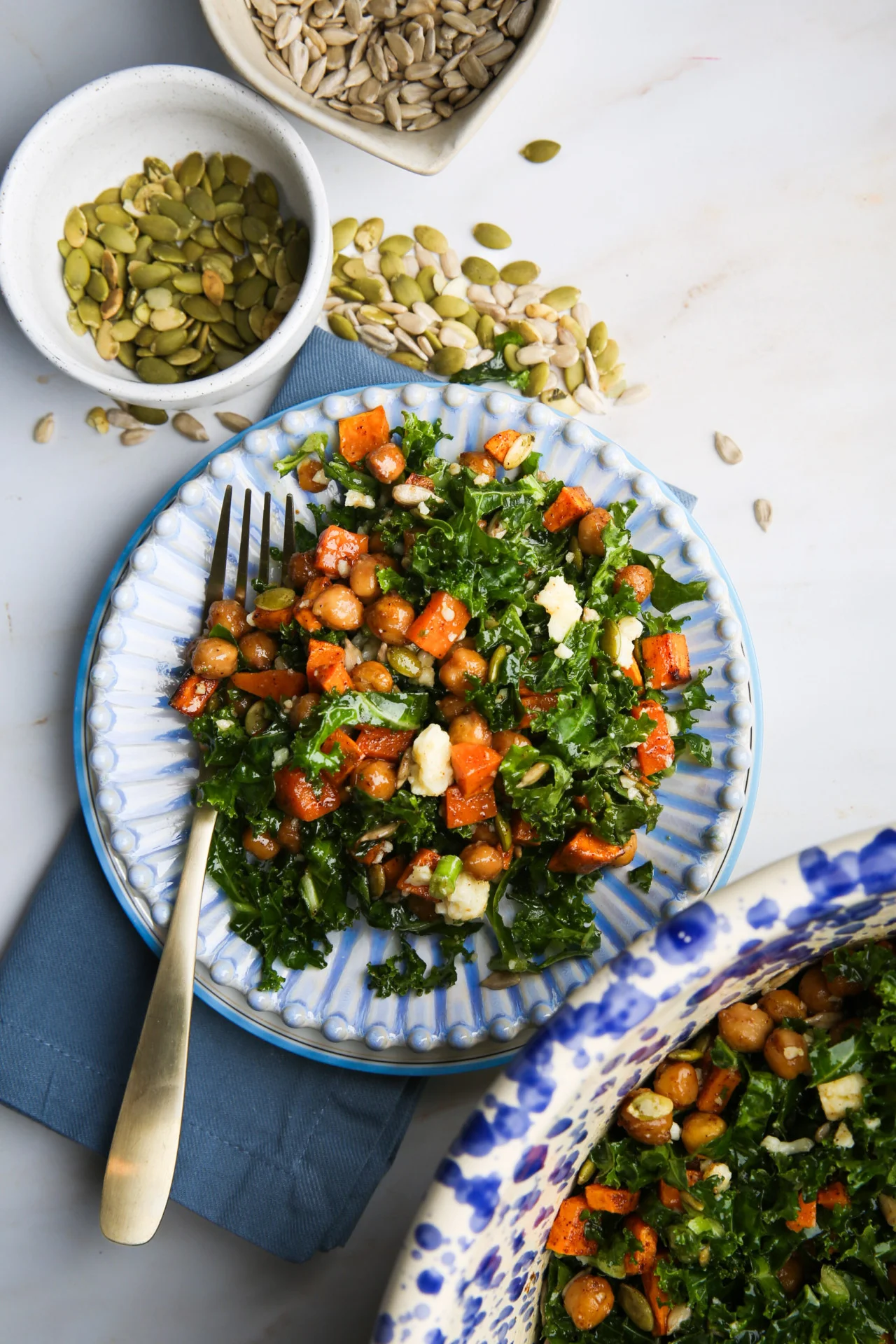 Una colorida ensalada de col rizada con boniatos asados, garbanzos y queso feta desmenuzado se rocía con aliño de Dijon al arce y se sirve en un plato azul decorativo. Sobre el plato, encima de una servilleta azul, hay un tenedor y, al lado, pequeños cuencos con semillas de calabaza y girasol. Se ve parcialmente un cuenco grande con más ensalada.